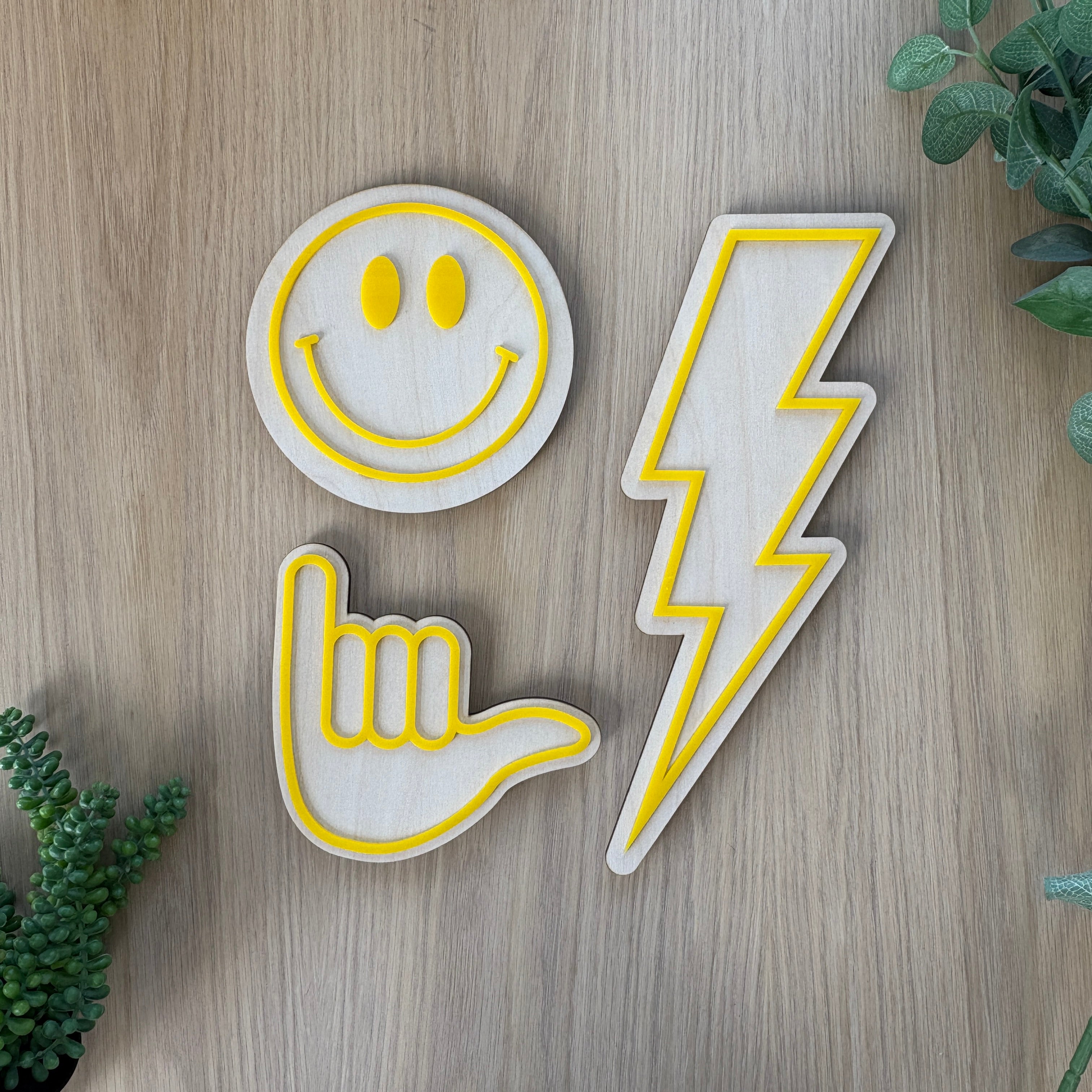 Three decorative patches shaped like a smiley face, hand sign, and lightning bolt on a wooden surface with greenery.