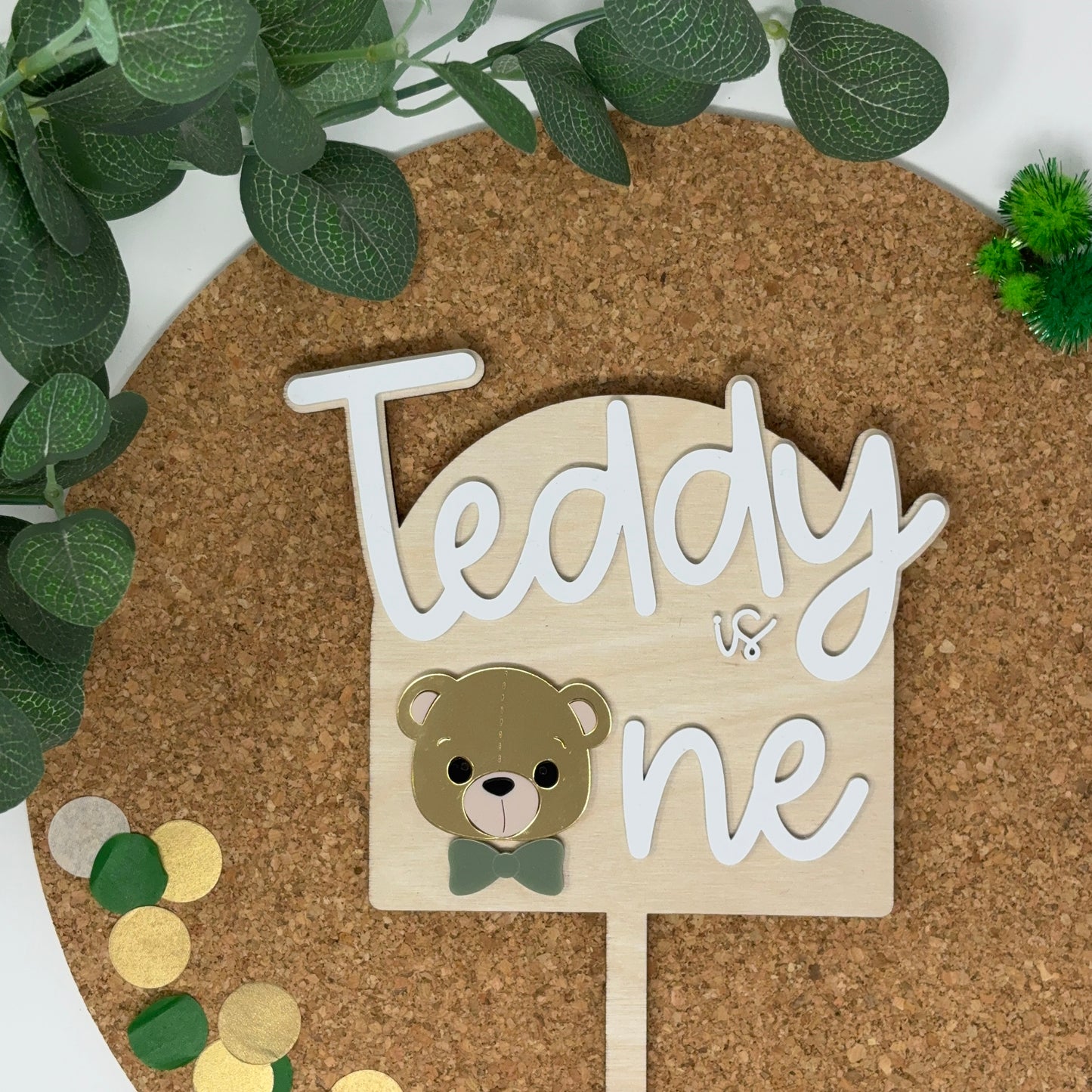 Wooden sign with 'Teddy is one' on a cork board with decorative elements.