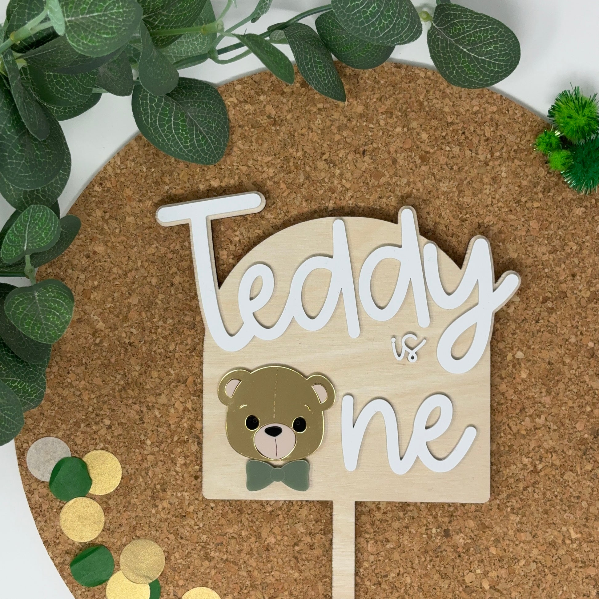 Wooden sign with 'Teddy is one' on a cork board with decorative elements.