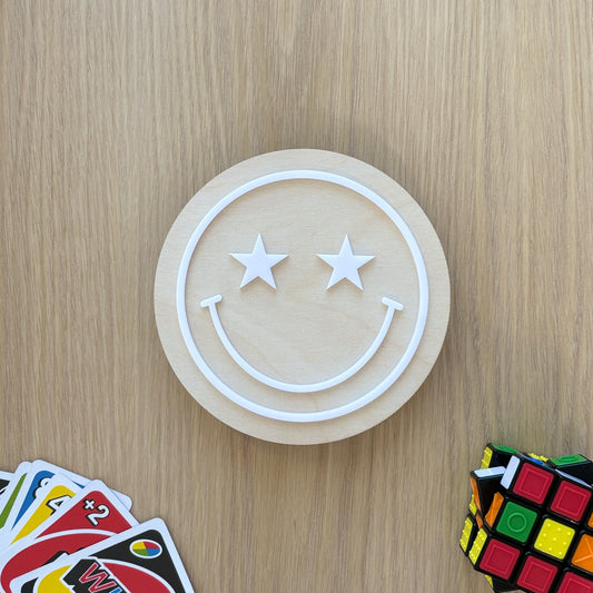 Circular wooden coaster with smiley face design on a wooden surface with cards and Rubik's cube.
