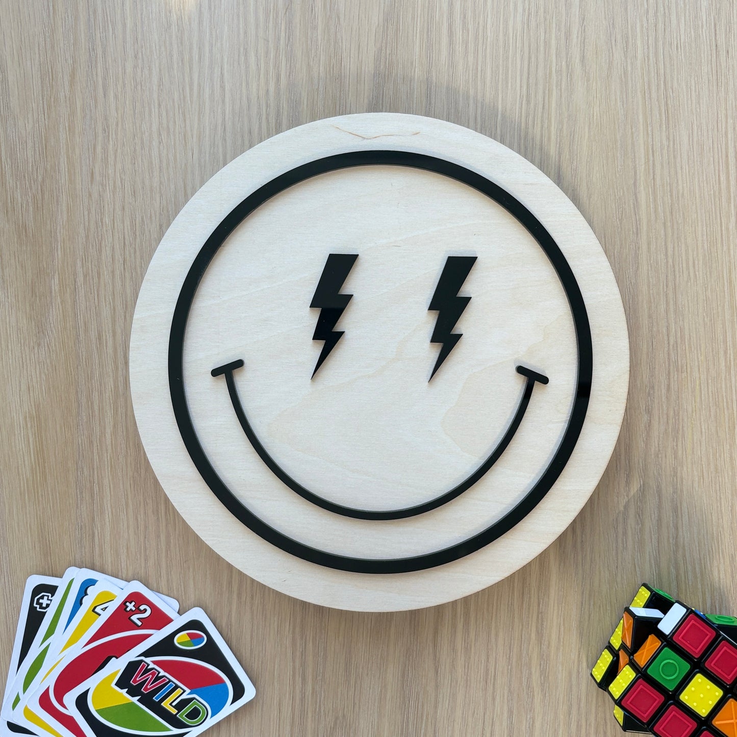 Smiley face design with lightning bolts on a wooden board, surrounded by cards and a Rubik's cube.