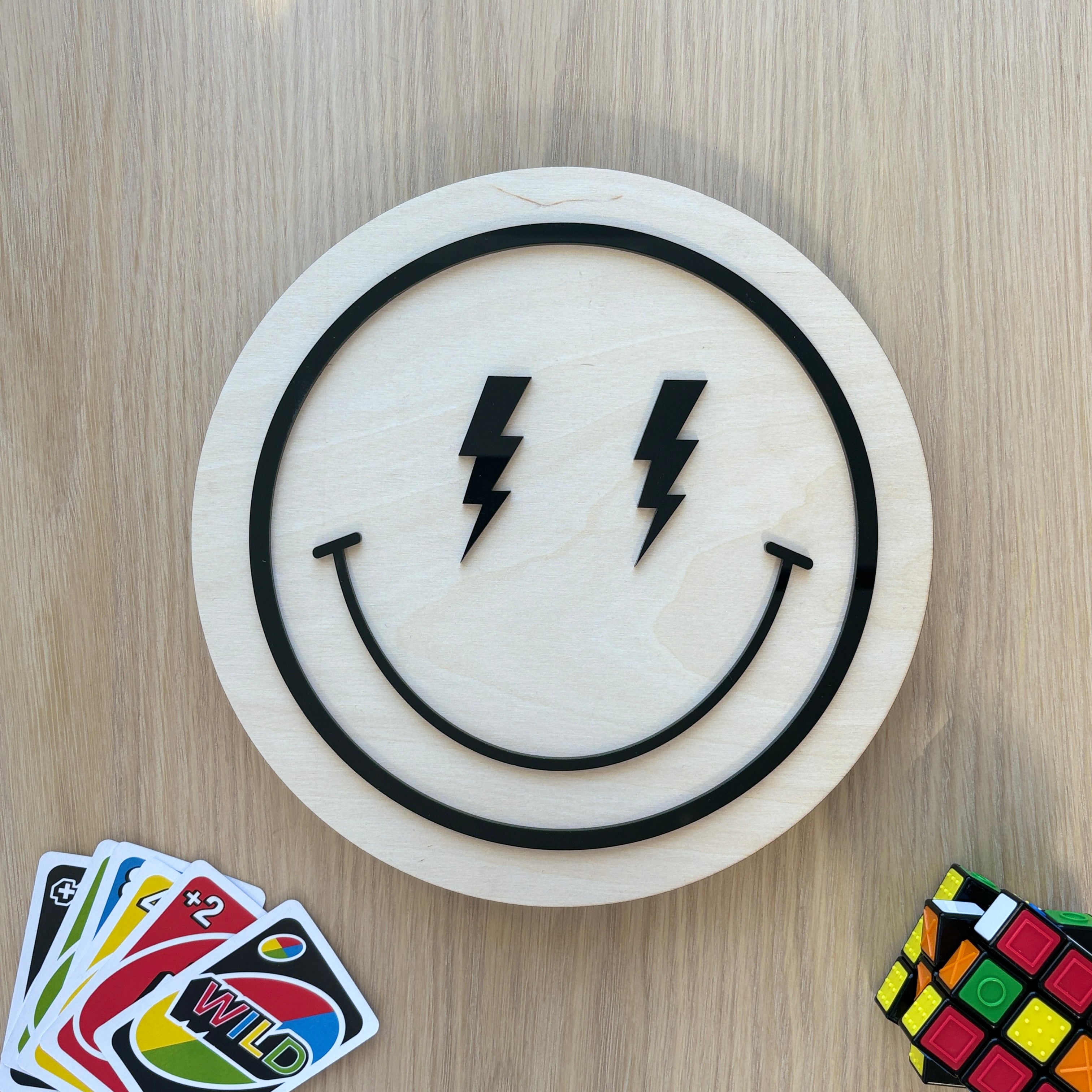 Smiley face design with lightning bolts on a wooden board, surrounded by cards and a Rubik's cube.