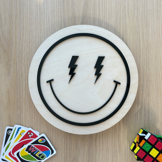 Smiley face design with lightning bolts on a wooden board, surrounded by cards and a Rubik's cube.