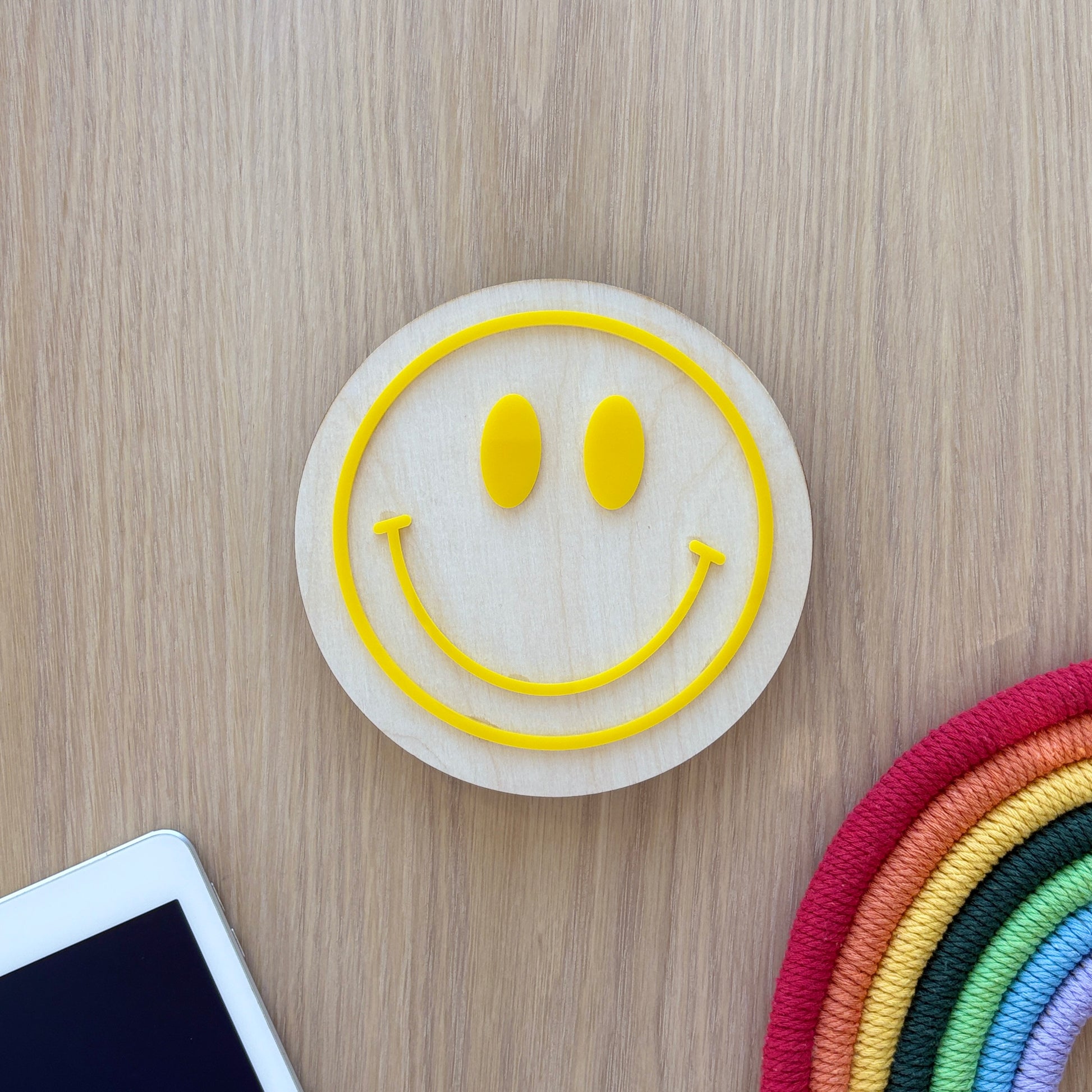 Smiley face coaster on a wooden surface with a smartphone and colorful woven item.