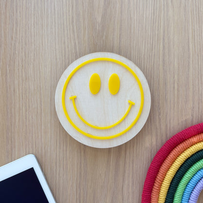 Smiley face coaster on a wooden surface with a smartphone and colorful woven item.