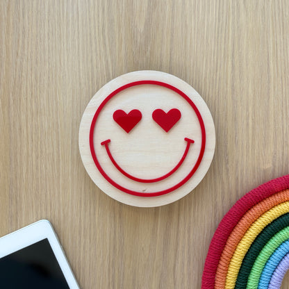 Smiley face with heart eyes on a wooden surface next to a smartphone and colorful object.