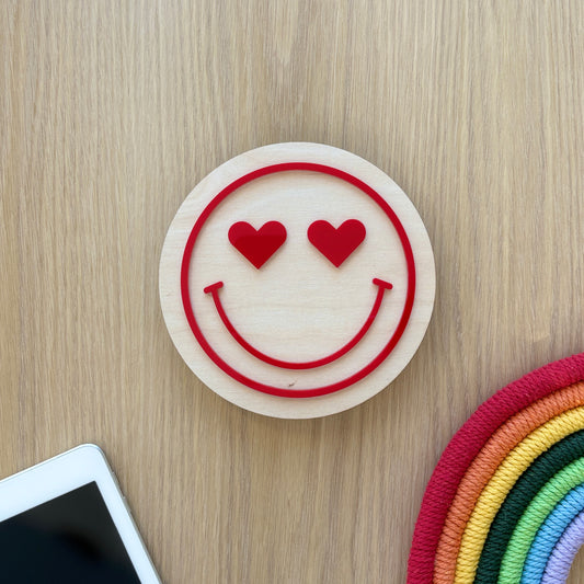 Smiley face with heart eyes on a wooden surface next to a smartphone and colorful object.