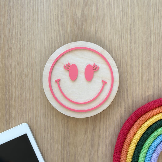 Smiley face coaster on a wooden surface with a smartphone and colorful woven item.