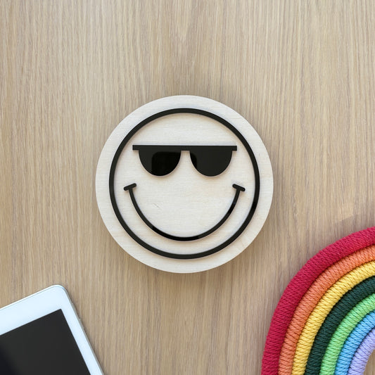 Smiley face with sunglasses on a wooden surface with a phone and colorful object.