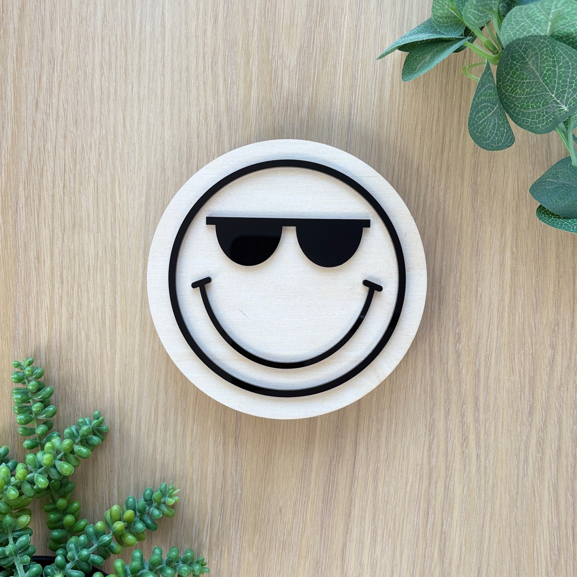 Round white coaster with smiley face design wearing sunglasses on a wooden surface with green plants.