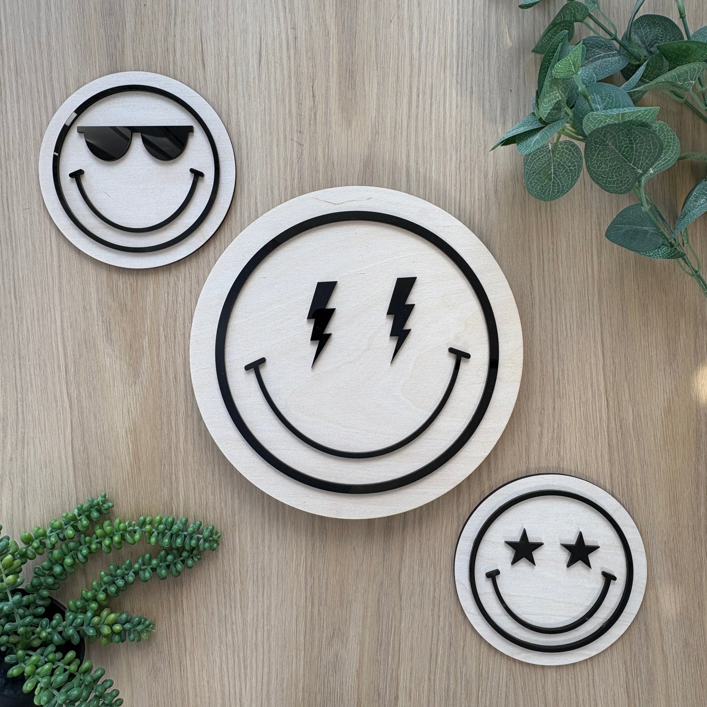 Three smiley face coasters with different designs on a wooden surface with plants.