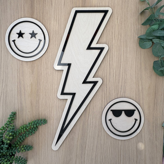 Three stickers on a wooden surface: a smiley face with stars, a lightning bolt, and a smiley face with sunglasses.
