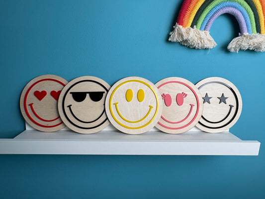 Five wooden coasters with smiley face designs on a white shelf against a blue wall with a rainbow decoration.